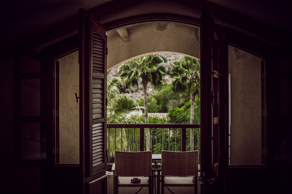 Interior of empty dark room with chairs near doorway on balcony with railing next to palms in daylight
