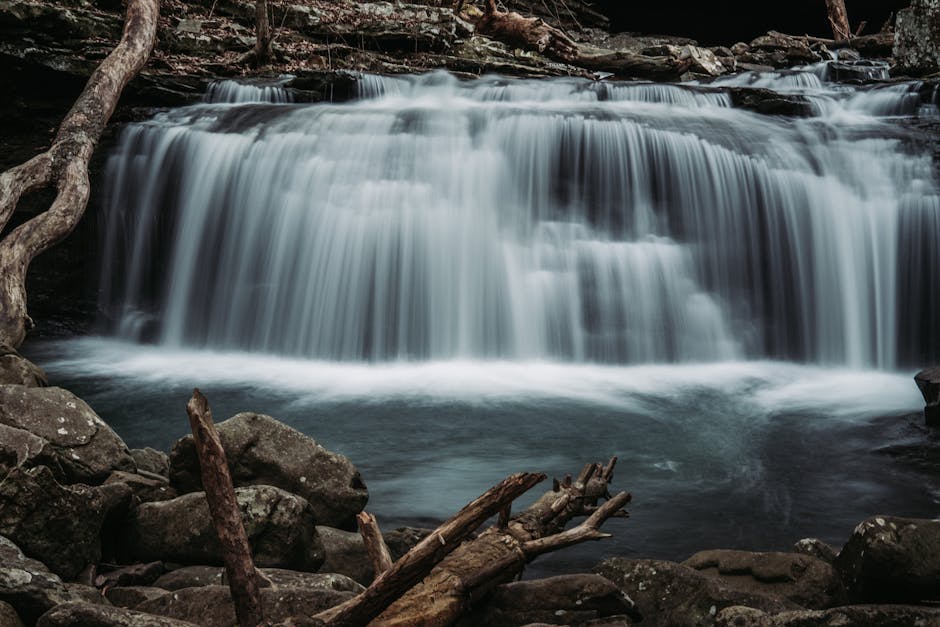 A serene view of a cascading waterfall in a forest, surrounded by rocks and logs, capturing the essence of nature.