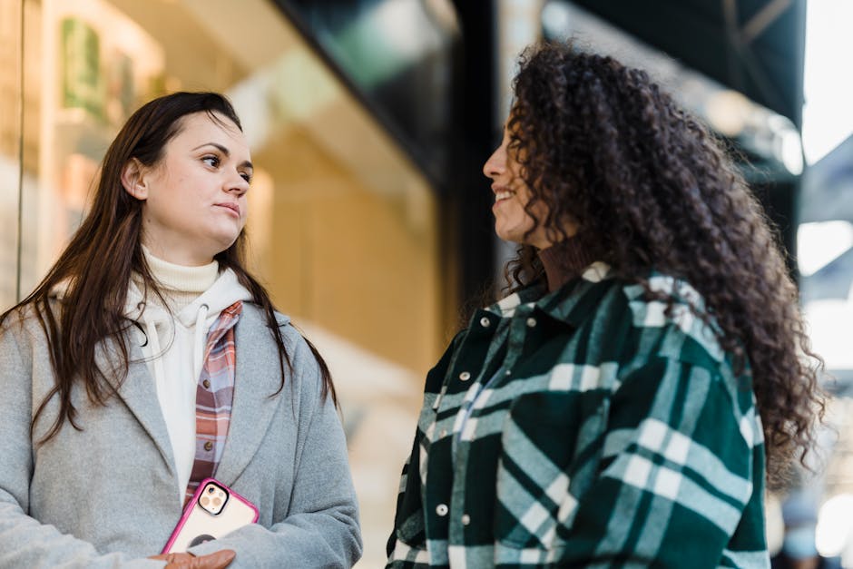 Side view of positive multiracial female friends looking at each other while standing near shop window on street on blurred background