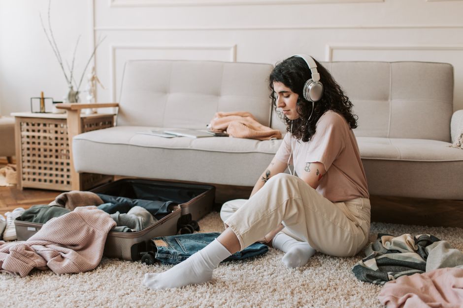 Young woman with headphones packing suitcase in a cozy living room.
