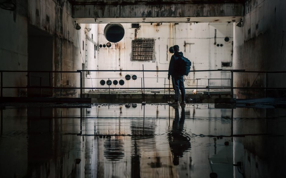 A person exploring a dark, abandoned industrial facility with water reflections.