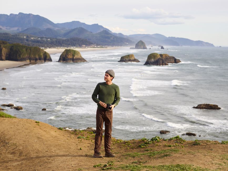 From above of attentive young male traveler with camera in casual clothes standing alone against rocky ocean and looking away