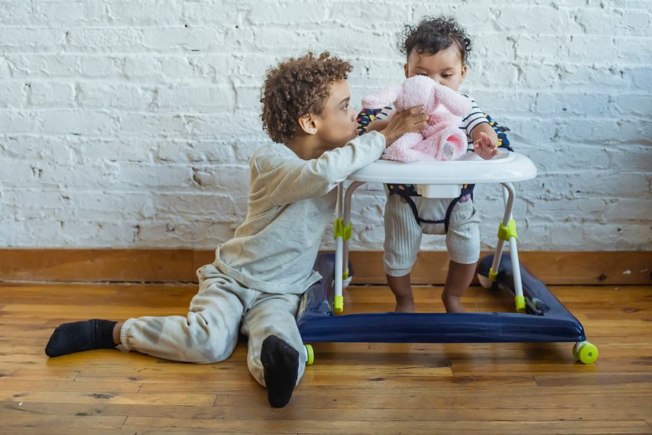 Full body of caring African American boy sitting on floor while playing with small toddler in baby walker near wall in light room