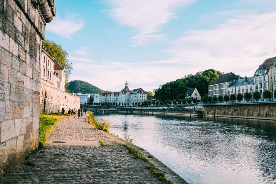 People walking along a scenic riverside path with historic architecture under a bright blue sky.