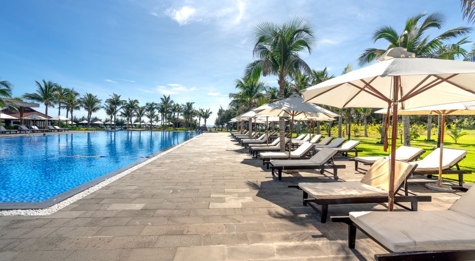 Beautiful tropical resort pool with sunloungers, parasols, and palm trees under a sunny sky.