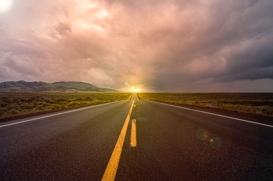 A scenic view of an empty highway stretching towards a sunset horizon under dramatic clouds.