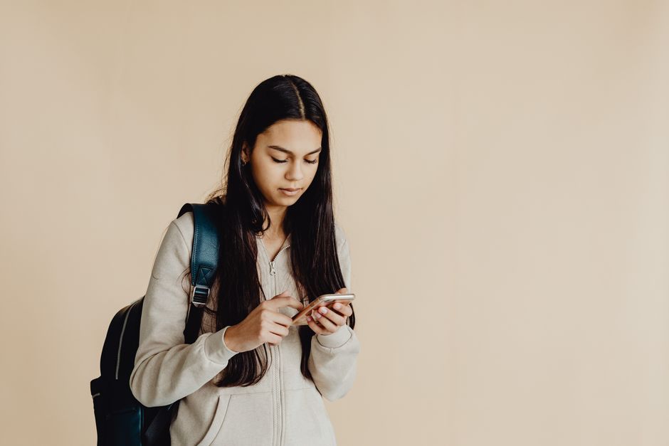 Teenage girl with backpack using a smartphone in a minimalist indoor setting, suited for educational themes.
