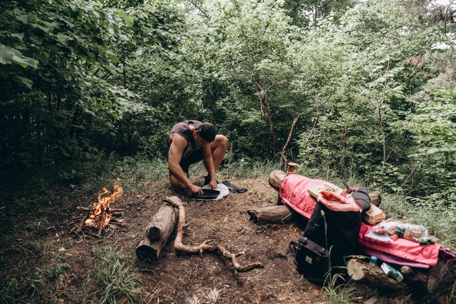Man setting up campfire in forest with gear spread out. Perfect for outdoor adventure themes.