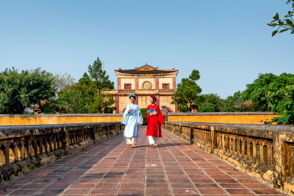 Two people in traditional Vietnamese clothing walk across a stone bridge towards a historic temple on a sunny day.