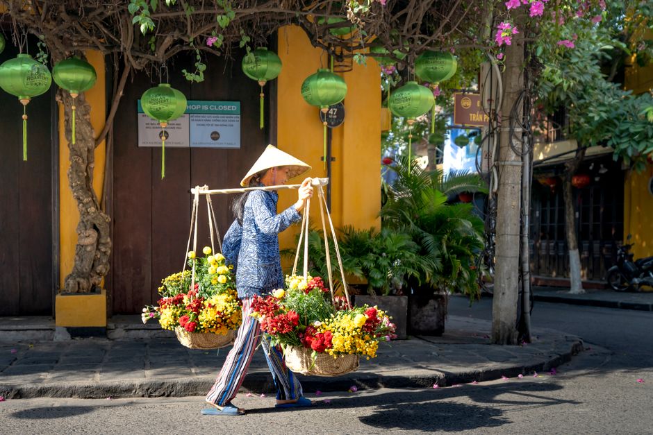 A Vietnamese woman in a conical hat carries colorful flowers on a street.