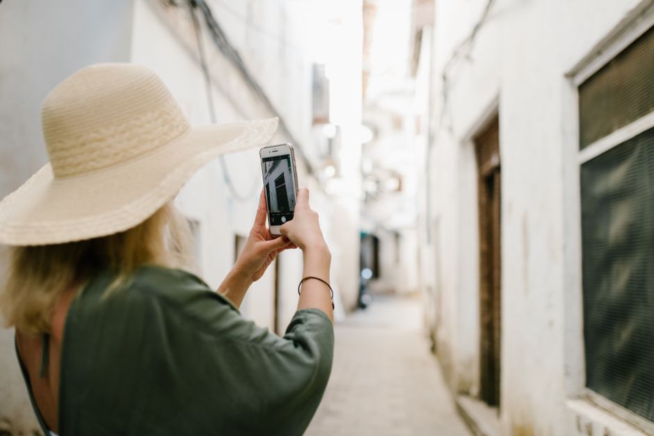 Back view of unrecognizable female tourist in hat taking photo on cellphone of old stone building while standing in narrow alley during trip