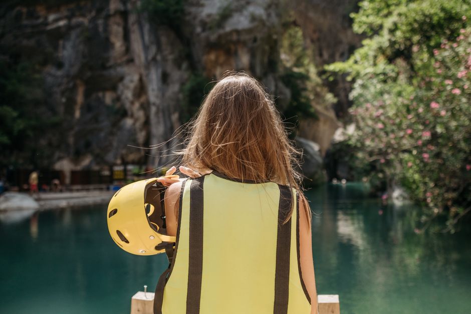 Young woman wearing a safety vest holding a helmet, overlooking a scenic water view with rocky cliffs.