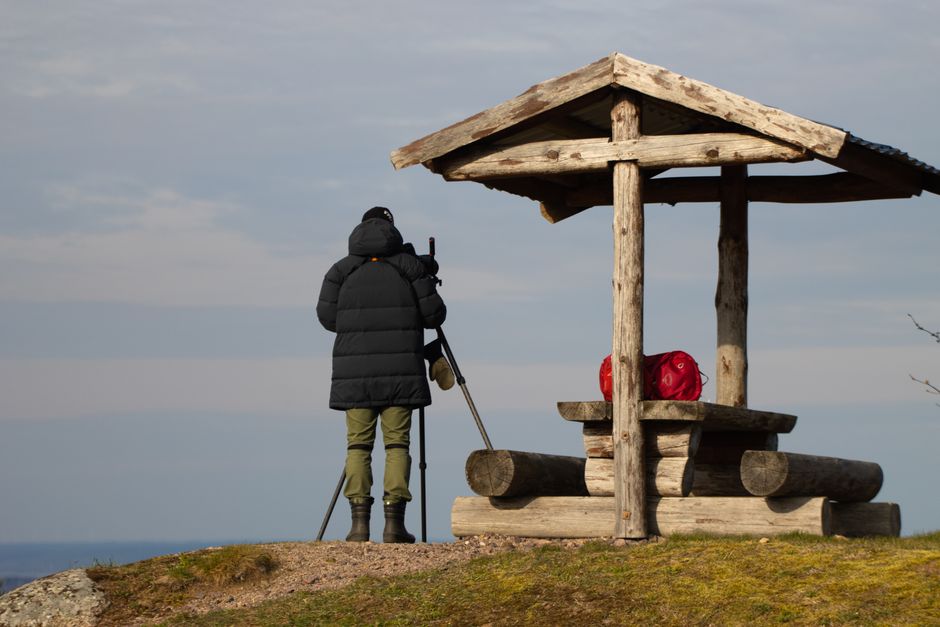 A hiker in winter gear takes photos from a scenic overlook in Sweden with a wooden shelter nearby.