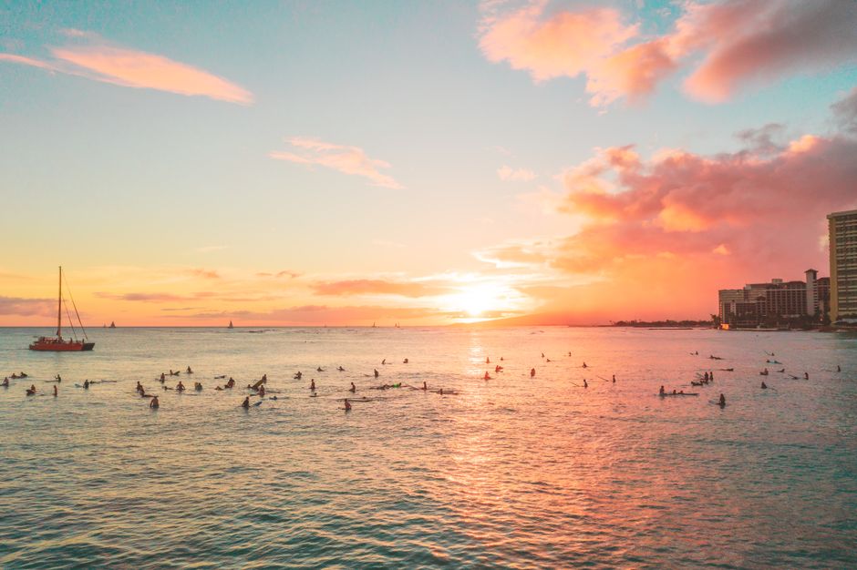 Surfers enjoy a vibrant sunset over Waikiki Beach, with sailboats on the horizon.