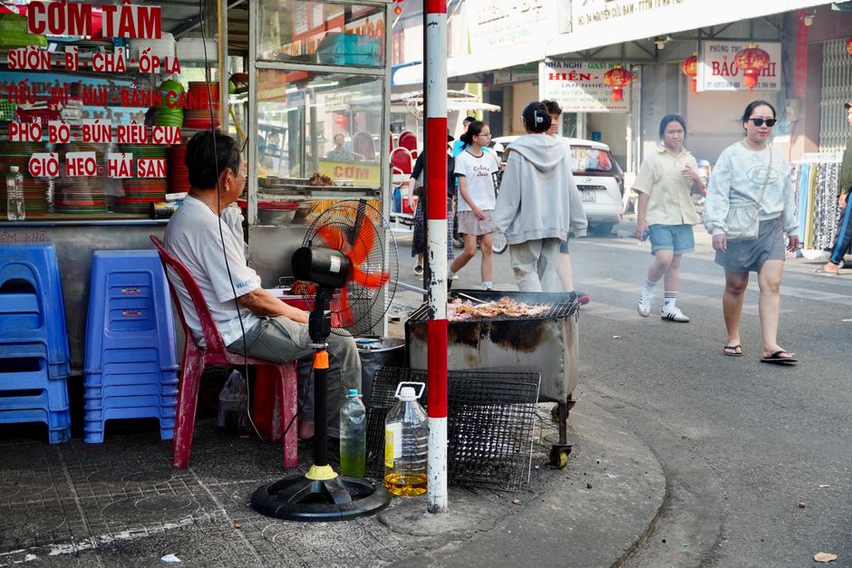 Grilling vendor at a bustling Ho Chi Minh City street with pedestrians.